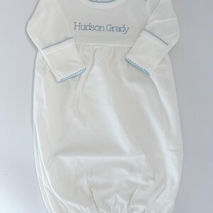 May include: A white long-sleeve baby gown featuring a round neckline with light blue scalloped trim. The cuffs also have matching light blue scalloped trim. The chest area is embroidered with the name Hudson Grady in light blue thread.