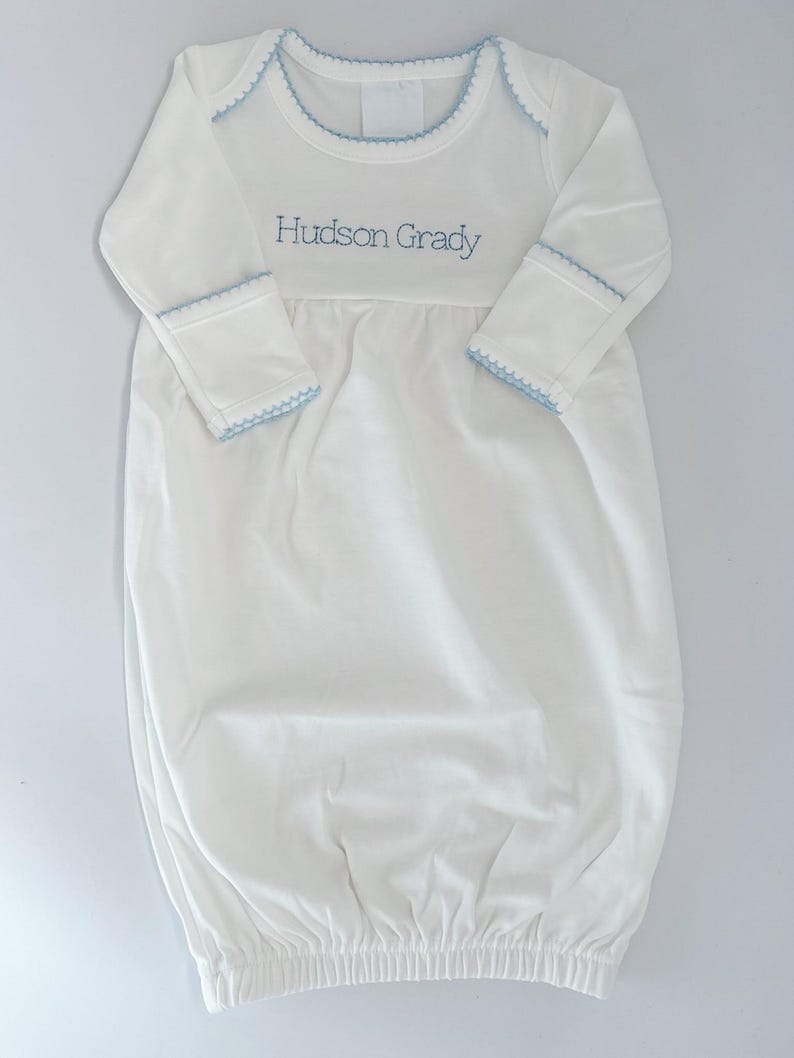 May include: A white long-sleeve baby gown featuring a round neckline with light blue scalloped trim. The cuffs also have matching light blue scalloped trim. The chest area is embroidered with the name Hudson Grady in light blue thread.