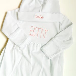 May include: A white long-sleeved gown and matching hat set. The hat features the name tate embroidered in red thread. The gown features the initials BTN embroidered in red thread on the chest. Both items have delicate white scalloped trim.