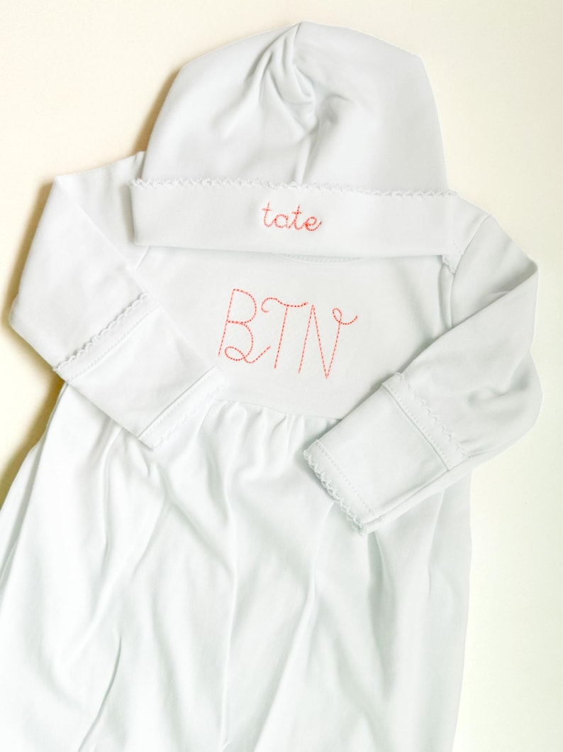 May include: A white long-sleeved gown and matching hat set. The hat features the name tate embroidered in red thread. The gown features the initials BTN embroidered in red thread on the chest. Both items have delicate white scalloped trim.