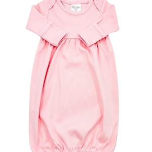 May include: A solid light pink long-sleeve baby gown featuring a round neckline with delicate scalloped trim, long sleeves with matching scalloped cuffs, and a gathered waistline that flows into a loose, open-bottom hem for easy access.