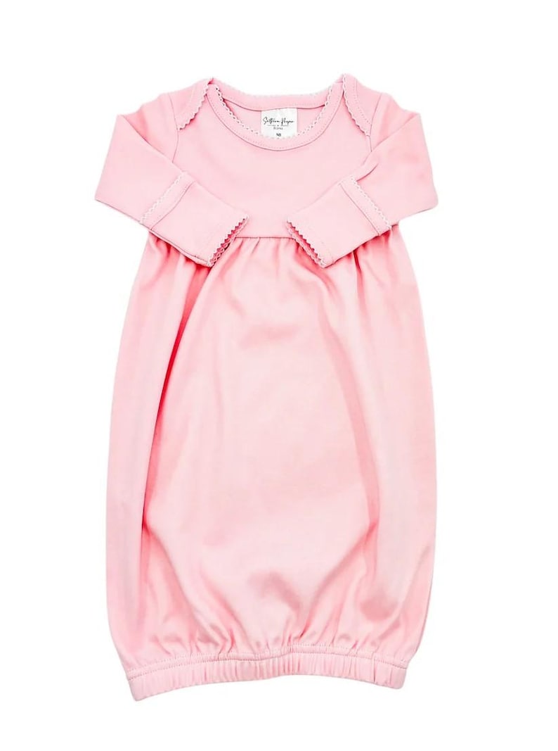 May include: A solid light pink long-sleeve baby gown featuring a round neckline with delicate scalloped trim, long sleeves with matching scalloped cuffs, and a gathered waistline that flows into a loose, open-bottom hem for easy access.