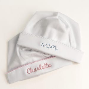 May include: Two white baby hats with embroidered names. One hat has "Sam" embroidered in blue, and the other has "Charlotte" embroidered in pink. Both hats have a scalloped edge.