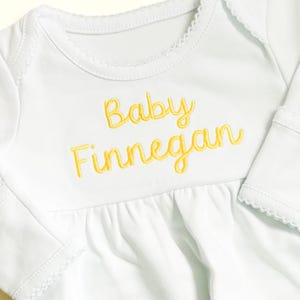 May include: A white long-sleeved baby bodysuit featuring the embroidered yellow text Baby Finnegan on the chest. The garment has a round neckline with delicate scalloped trim along the collar and sleeve cuffs, and a gathered waistline detail.