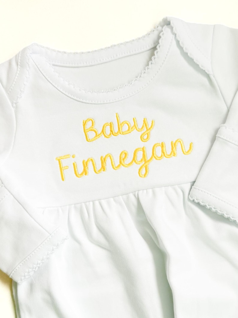 May include: A white long-sleeved baby bodysuit featuring the embroidered yellow text Baby Finnegan on the chest. The garment has a round neckline with delicate scalloped trim along the collar and sleeve cuffs, and a gathered waistline detail.