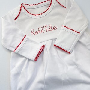 May include: A white long-sleeve infant gown featuring red scalloped trim along the neckline and sleeve cuffs. The chest area is embroidered with the red text Roll Tide in a cursive script font, and the garment has a gathered bodice design.
