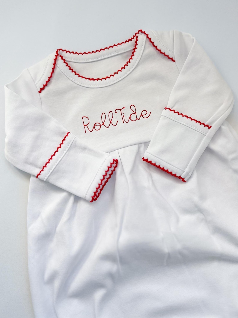 May include: A white long-sleeve infant gown featuring red scalloped trim along the neckline and sleeve cuffs. The chest area is embroidered with the red text Roll Tide in a cursive script font, and the garment has a gathered bodice design.