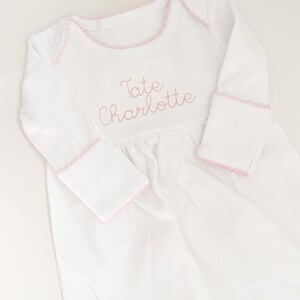 May include: A white long-sleeved baby gown featuring a round neckline with pink picot trim. The chest is embroidered with the name Tate Charlotte in pink thread. The sleeves have matching pink picot trim at the cuffs and a gathered waistline.