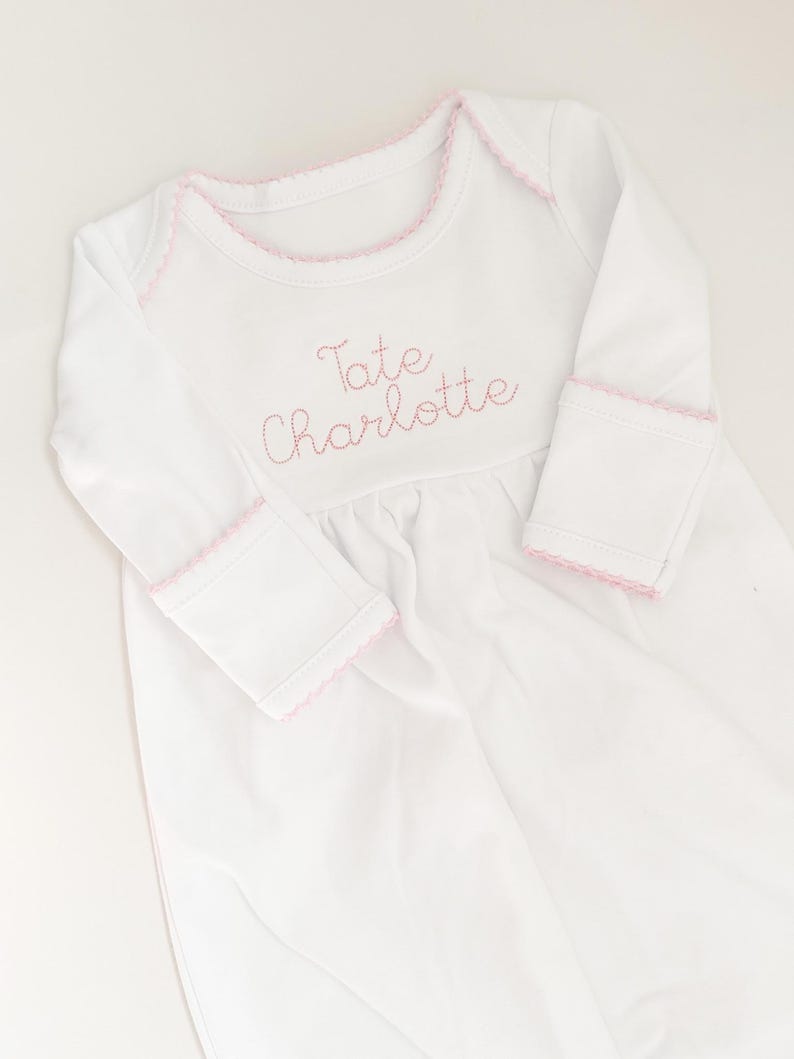 May include: A white long-sleeved baby gown featuring a round neckline with pink picot trim. The chest is embroidered with the name Tate Charlotte in pink thread. The sleeves have matching pink picot trim at the cuffs and a gathered waistline.