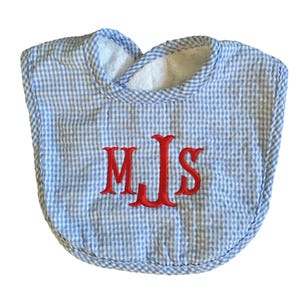 May include: A baby bib with a blue and white gingham pattern. The bib has a white terry cloth lining around the neck and is embroidered with the red initials "MJS".