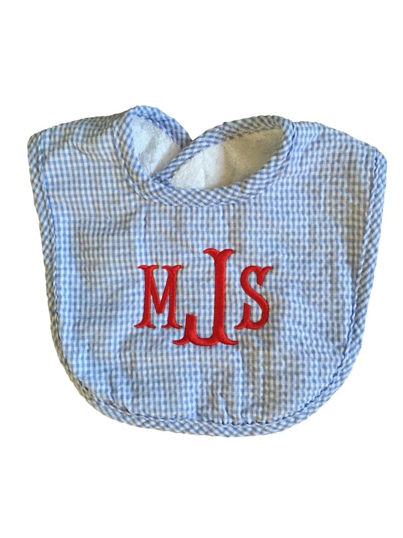 May include: A baby bib with a blue and white gingham pattern. The bib has a white terry cloth lining around the neck and is embroidered with the red initials "MJS".