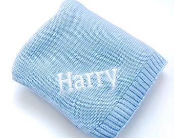 May include: A light blue knitted blanket featuring a ribbed border and the name Harry embroidered in white thread across the center. The textile has a consistent garter stitch pattern throughout the main body of the folded fabric.