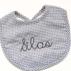 May include: A gray and white gingham baby bib with the name "Silas" embroidered in black cursive script. The bib has a rounded shape and a soft, padded texture, perfect for catching spills.