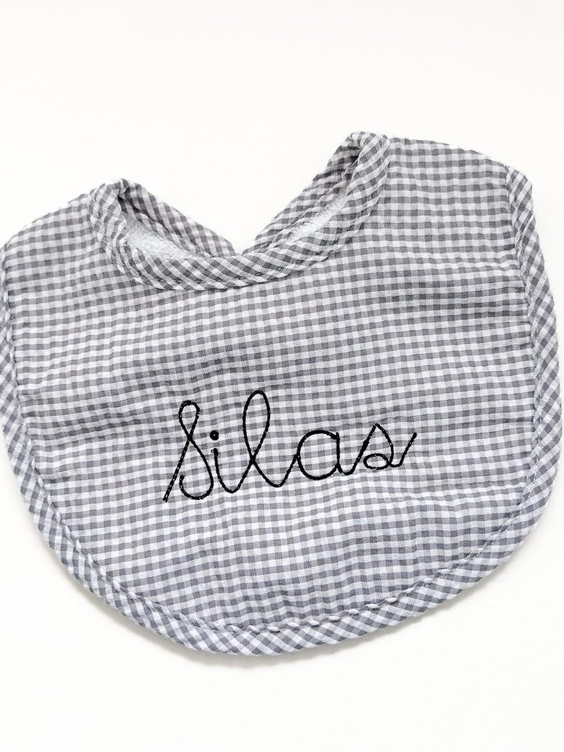 May include: A gray and white gingham baby bib with the name "Silas" embroidered in black cursive script. The bib has a rounded shape and a soft, padded texture, perfect for catching spills.
