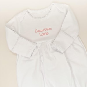 May include: A white long-sleeved baby gown featuring a round neckline with delicate scalloped trim. The chest area is embroidered with the name Dawson Lane in soft pink cursive lettering. The sleeves have matching scalloped trim at the cuffs.