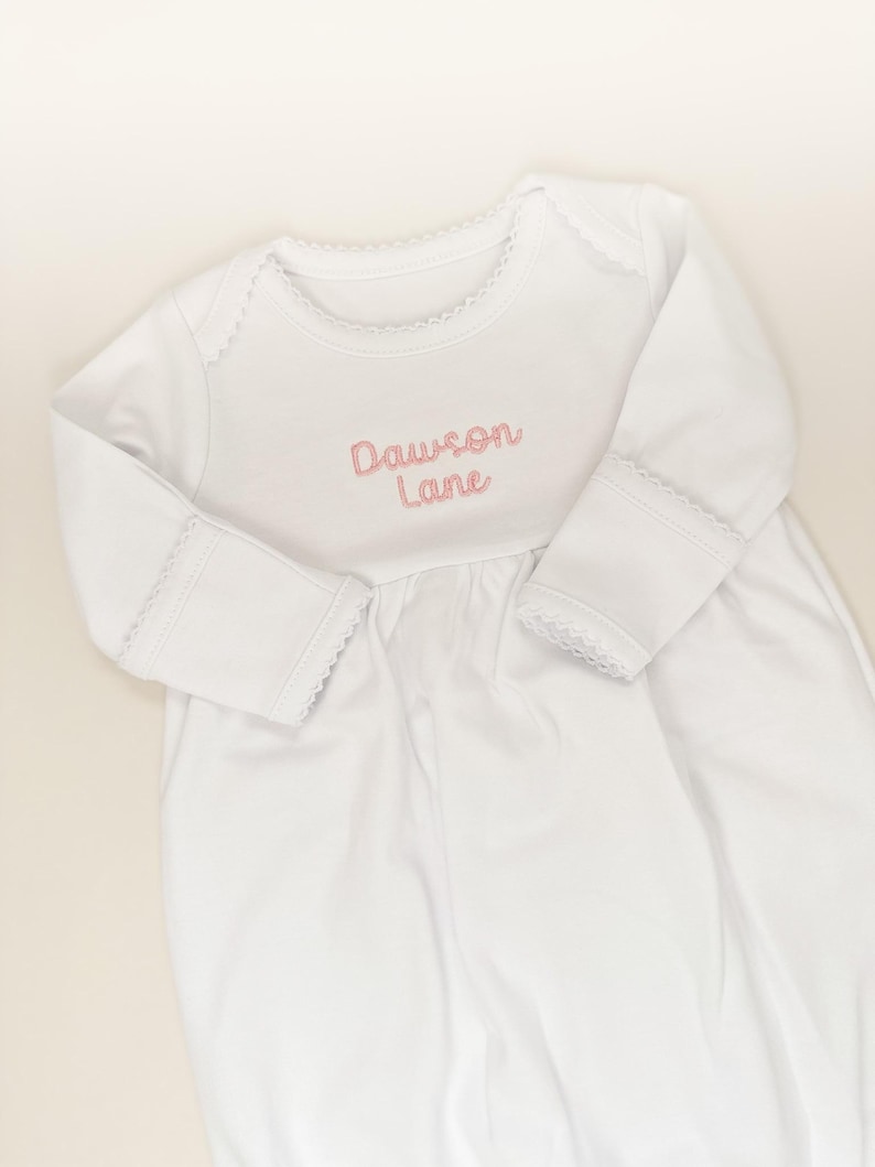 May include: A white long-sleeved baby gown featuring a round neckline with delicate scalloped trim. The chest area is embroidered with the name Dawson Lane in soft pink cursive lettering. The sleeves have matching scalloped trim at the cuffs.