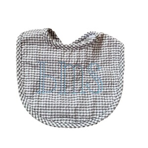 May include: A gray and white gingham baby bib with the name "BILL" embroidered in blue thread. The bib has a rounded shape and a soft, quilted appearance, perfect for catching spills.