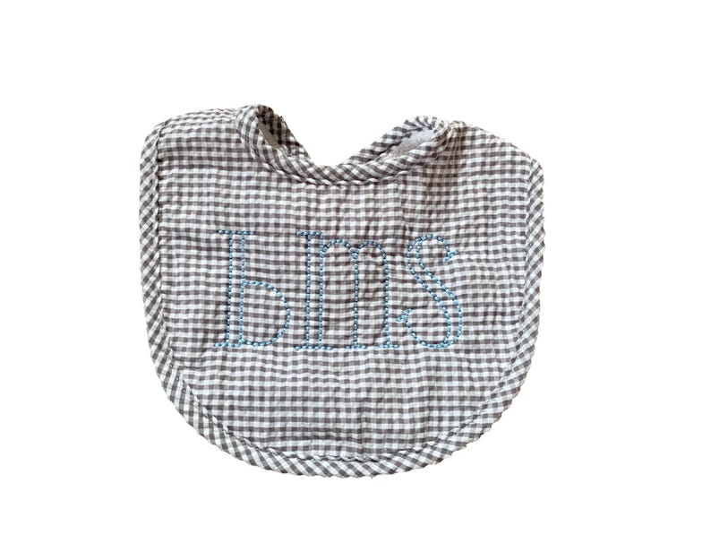 May include: A gray and white gingham baby bib with the name "BILL" embroidered in blue thread. The bib has a rounded shape and a soft, quilted appearance, perfect for catching spills.