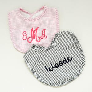 May include: Two baby bibs. One pink gingham bib with the embroidered letters "SMA" in pink. The other is a gray and white gingham bib with the embroidered name "Woods" in black. Both bibs have a rounded shape.
