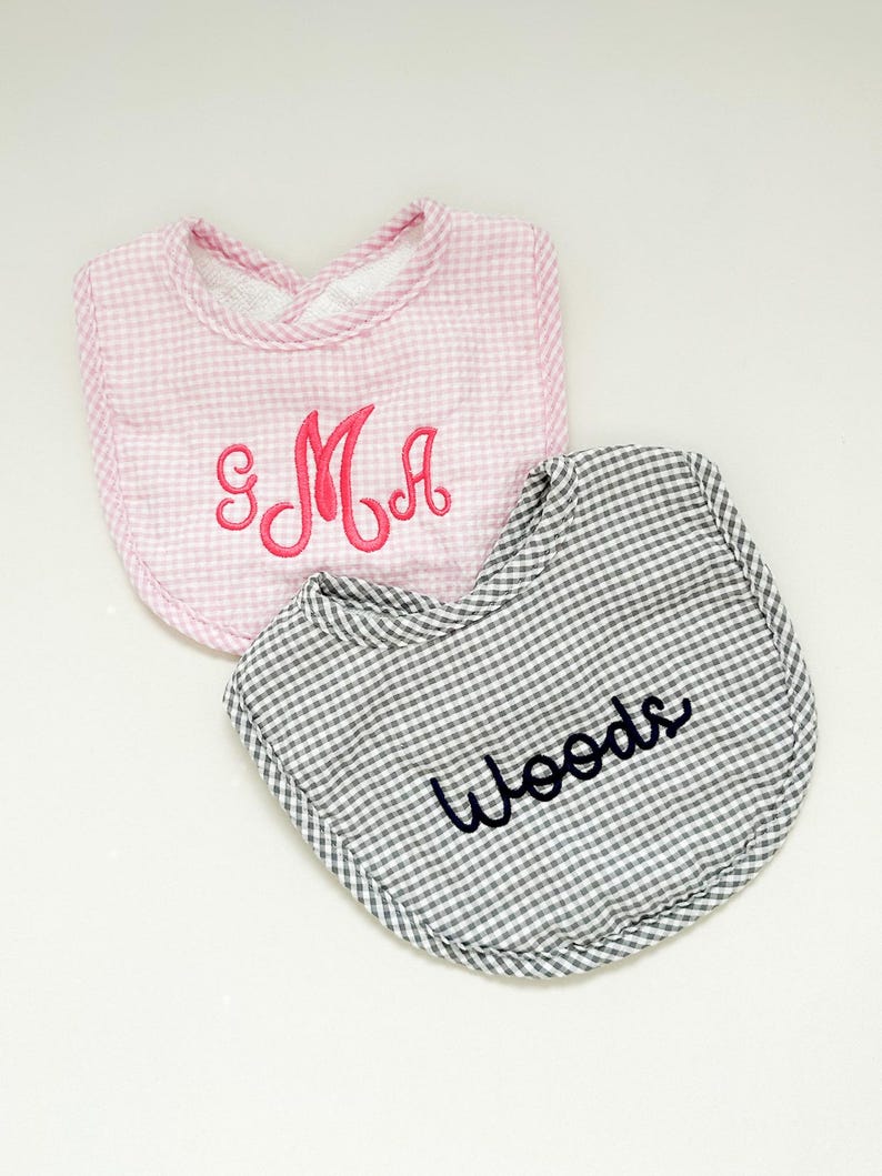 May include: Two baby bibs. One pink gingham bib with the embroidered letters "SMA" in pink. The other is a gray and white gingham bib with the embroidered name "Woods" in black. Both bibs have a rounded shape.