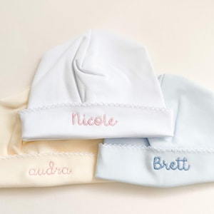 May include: Three baby hats in cream, white, and light blue. Each hat has a scalloped brim and embroidered name: "Audra", "Nicole", and "Brett". The hats are soft and appear to be made of a lightweight fabric, perfect for newborns.