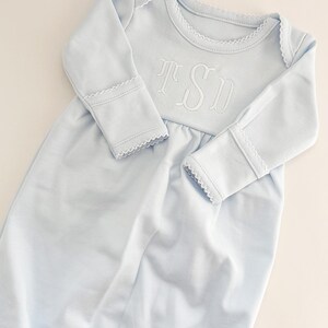 May include: A light blue long-sleeved baby gown featuring a white embroidered monogram with the letters TSD on the chest. The garment has a round neckline and sleeve cuffs finished with a delicate white scalloped picot trim detail.