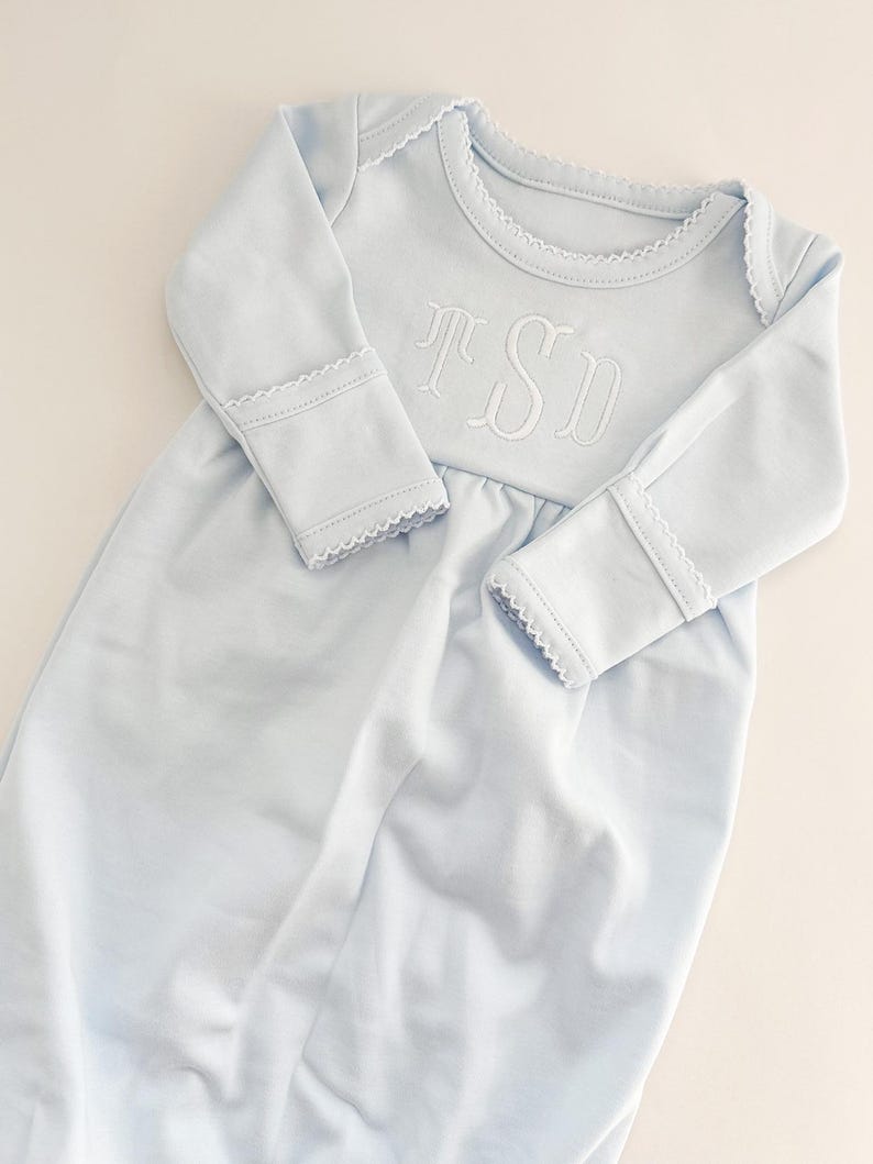 May include: A light blue long-sleeved baby gown featuring a white embroidered monogram with the letters TSD on the chest. The garment has a round neckline and sleeve cuffs finished with a delicate white scalloped picot trim detail.