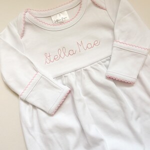 May include: A white long-sleeved baby gown featuring pink picot trim along the neckline and sleeve cuffs. The chest area is embroidered with the name Stella Mae in pink thread. A brand label reading Southern Proper is visible at the neckline.