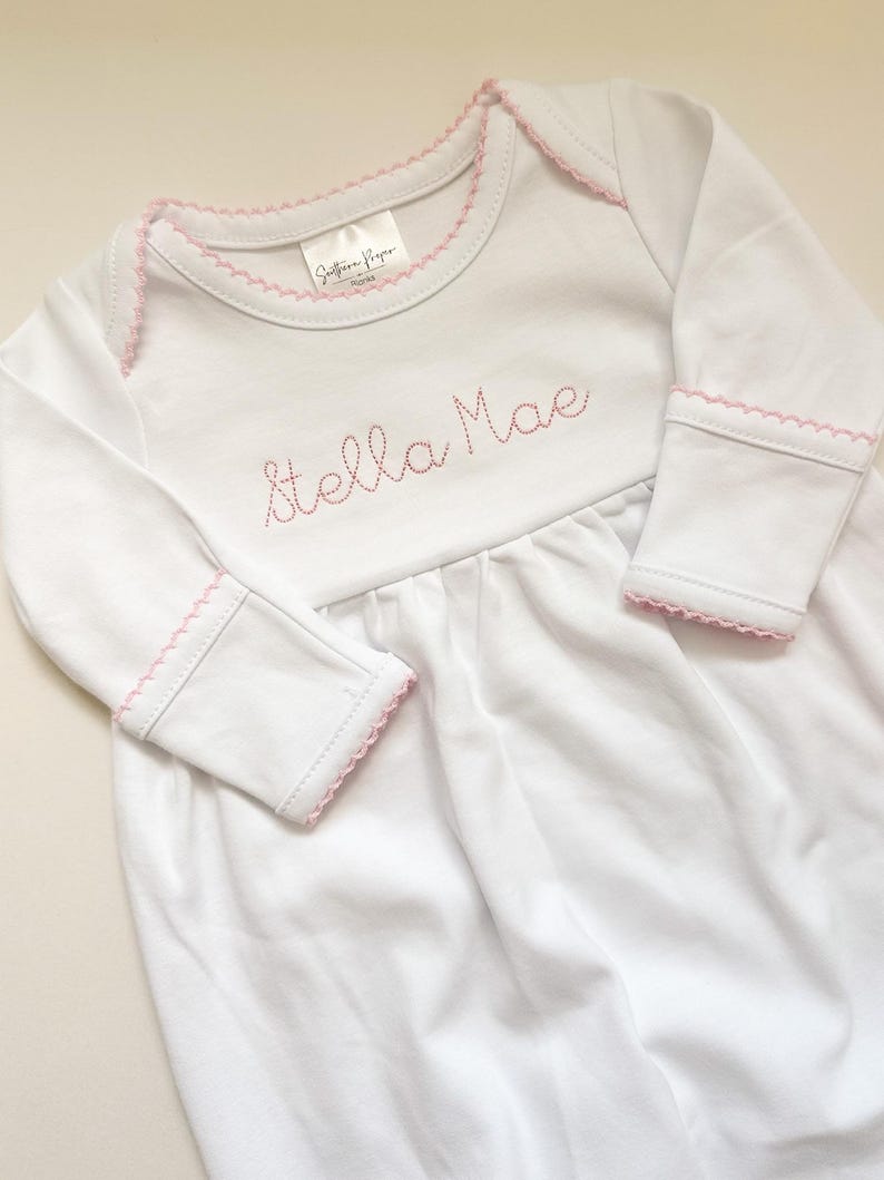 May include: A white long-sleeved baby gown featuring pink picot trim along the neckline and sleeve cuffs. The chest area is embroidered with the name Stella Mae in pink thread. A brand label reading Southern Proper is visible at the neckline.