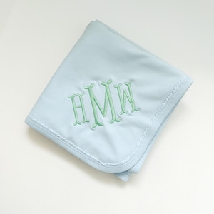 May include: A light blue baby blanket featuring a decorative scalloped edge and a centered monogram embroidered in light green thread. The monogram consists of the letters H, M, and W in a stylized, ornate font, centered on the fabric.