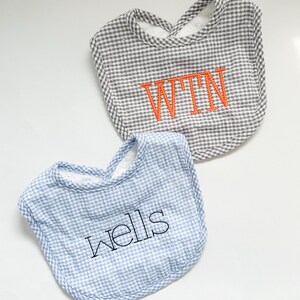 May include: Two baby bibs. One is gray and white gingham with orange embroidered initials "WTN". The other is blue and white gingham with the name "wells" embroidered in black. Both have a soft, white terrycloth lining.