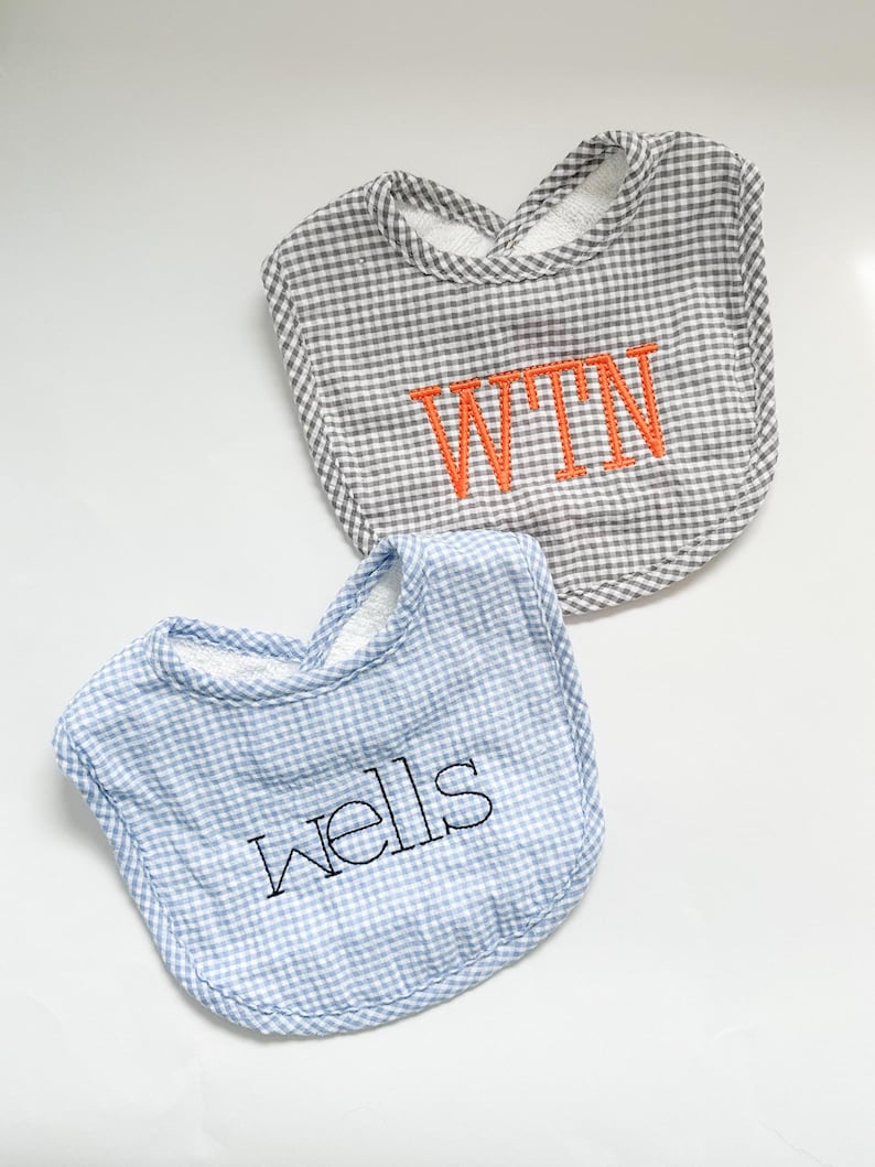 May include: Two baby bibs. One is gray and white gingham with orange embroidered initials "WTN". The other is blue and white gingham with the name "wells" embroidered in black. Both have a soft, white terrycloth lining.
