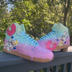 May include: Custom high-top sneakers with a colorful, whimsical design. The shoes feature a gradient of pink, blue, and green, with a brown sole and a dripping effect. The design includes donuts and cartoon astronauts, with sprinkles scattered across the surface.