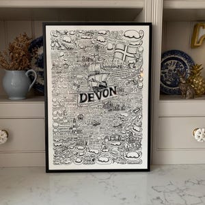 May include: A black and white illustrated map of Devon, England, with the text "Devon" in a large font at the bottom. The map includes various landmarks, towns, and cities, including Plymouth, Exeter, and Dartmoor National Park. The map is framed in a black frame and is hanging on a white wall.