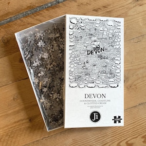 May include: A 1000-piece jigsaw puzzle box with a black and white illustration of Devon's countryside and coastline. The box reads "DEVON" in large letters. The puzzle pieces are visible in a clear plastic bag, ready to be assembled.