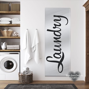 May include: A black and white vinyl wall decal with the word "Laundry" written in a cursive font on a silver background. The decal is mounted on a white wall in a bathroom setting.