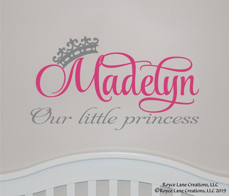 K&ouml;nnte beinhalten: Wandaufkleber mit dem Namen "Madelyn" in rosa Schrift, gekr&ouml;nt von einer grauen Krone. Darunter steht der Satz "Our little princess" in Grau. Das Design befindet sich auf einem hellgrauen Hintergrund.