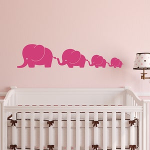 Peut inclure: Un autocollant mural de cr&egrave;che rose repr&eacute;sentant une ligne de cinq &eacute;l&eacute;phants de diff&eacute;rentes tailles, chacun avec une trompe lev&eacute;e et reli&eacute;e &agrave; celle qui le pr&eacute;c&egrave;de. Les &eacute;l&eacute;phants sont sur un mur rose, avec un berceau blanc et une lampe en arri&egrave;re-plan.