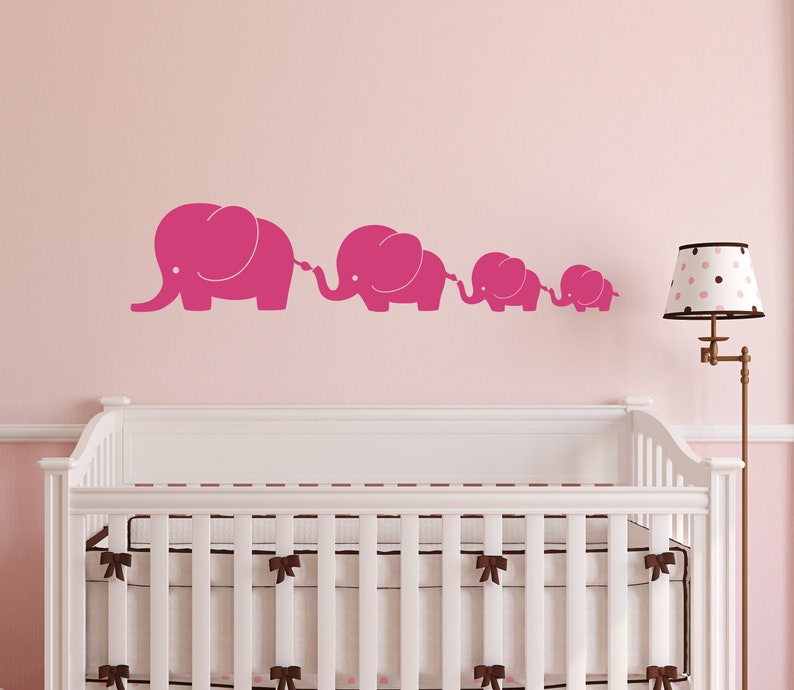 Peut inclure: Un autocollant mural de cr&egrave;che rose repr&eacute;sentant une ligne de cinq &eacute;l&eacute;phants de diff&eacute;rentes tailles, chacun avec une trompe lev&eacute;e et reli&eacute;e &agrave; celle qui le pr&eacute;c&egrave;de. Les &eacute;l&eacute;phants sont sur un mur rose, avec un berceau blanc et une lampe en arri&egrave;re-plan.