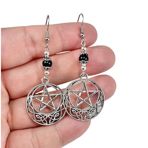 May include: A pair of silver-toned dangle earrings featuring a circular pendant with an intricate pentagram design and Celtic knotwork. Each earring is accented with a single round black bead and a silver-toned metal hook for pierced ears.