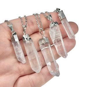May include: Five clear quartz crystal point pendants suspended on silver-toned metal chains. Each natural stone features a unique, elongated hexagonal shape with a metallic cap at the top, showcasing varying degrees of transparency and internal inclusions.