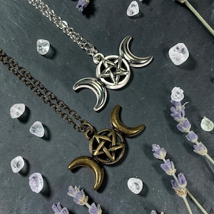 May include: Two necklaces featuring a triple moon goddess symbol with a central pentagram. One pendant is silver-toned on a silver chain, and the other is bronze-toned on a matching bronze chain. Both are displayed with small clear crystals and lavender.