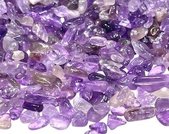 May include: Close-up of a pile of amethyst gemstone chips. The chips vary in size and shape, displaying shades of purple, lavender, and translucent white. The gemstones are scattered, creating a textured surface.