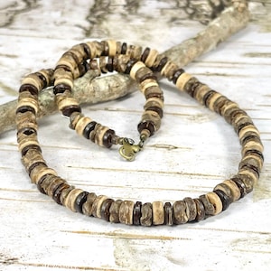 May include: A handcrafted necklace made of natural, round wooden beads in shades of brown and tan. The beads are strung together with a metal clasp. The necklace is displayed on a light-colored wooden surface.