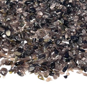 May include: Close-up of a pile of polished, crushed gemstones. The stones are a mix of dark gray, black, and translucent smoky quartz, with a glossy finish. The irregular shapes and sizes create a textured surface.