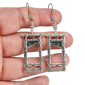 May include: A pair of silver guillotine earrings. The earrings are shaped like a guillotine with a blade and a wooden frame. The earrings are hanging from silver hooks.