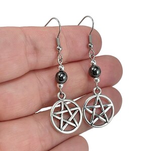 May include: A pair of silver-toned dangle earrings featuring a circular pentagram charm suspended from a single dark gray, metallic-looking spherical bead. The earrings are attached to standard silver-toned metal fishhook ear wires.
