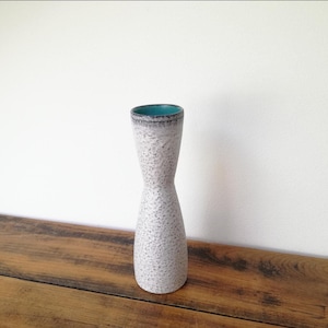 May include: A slender, ceramic vase with a distinctive hourglass silhouette. The vase has a speckled, off-white finish and a teal interior. It stands on a wooden surface.