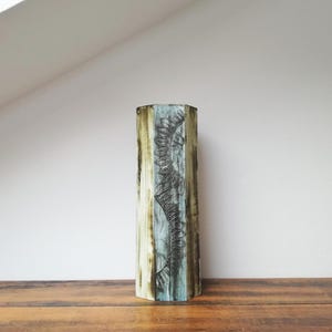 May include: A tall, hexagonal vase with a textured surface. The vase features a vertical, stylized design in black against a backdrop of muted blue and tan hues. The vase is displayed on a wooden surface.