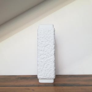 Könnte beinhalten: Eine hohe, rechteckige weiße Keramikvase mit strukturierter Oberfläche. Die Vase hat einen quadratischen Sockel und eine etwas breitere Oberseite. Sie steht auf einer dunklen Holzoberfläche vor einer weißen Wand.